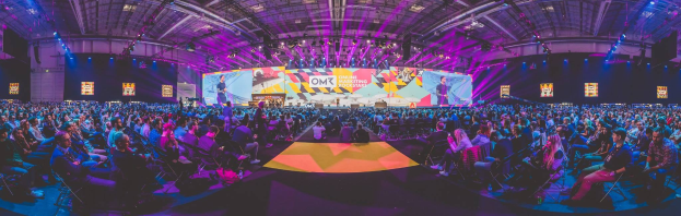 Eine große Menschenmenge sitzt auf Stühlen, die einer hell erleuchteten Bühne mit Bildschirmen zugewandt sind, auf der mehrere Personen stehen.