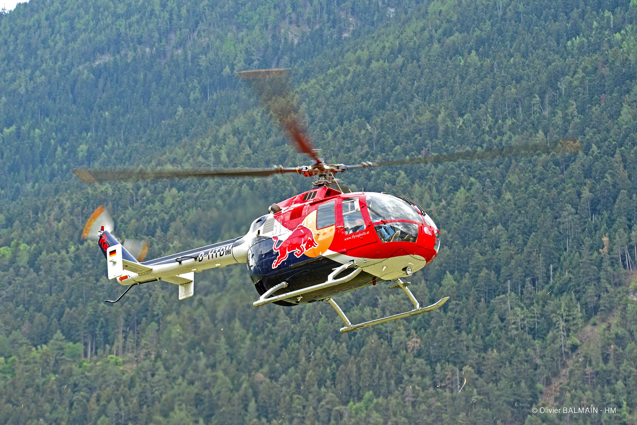 Roter Bull-Hubschrauber fliegt über einen grünen Wald mit Bäumen auf Hügeln und einem klaren blauen Himmel.
