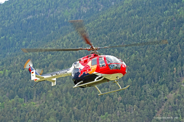 Roter Bull-Hubschrauber fliegt über einen grünen Wald mit Bäumen auf Hügeln und einem klaren blauen Himmel.