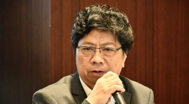 Ein Mann im Anzug mit einem Mikrofon, der leidenschaftlich zu sprechen scheint, vor einer hölzernen Wand.