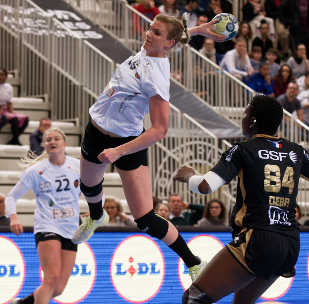 Eine Gruppe von Frauen, die Handball auf einem Platz mit einem Ball in der Luft spielen, Zuschauer im Hintergrund und ein "Handball-Weltmeisterschaft 2019 - Tag 2"-Schild.