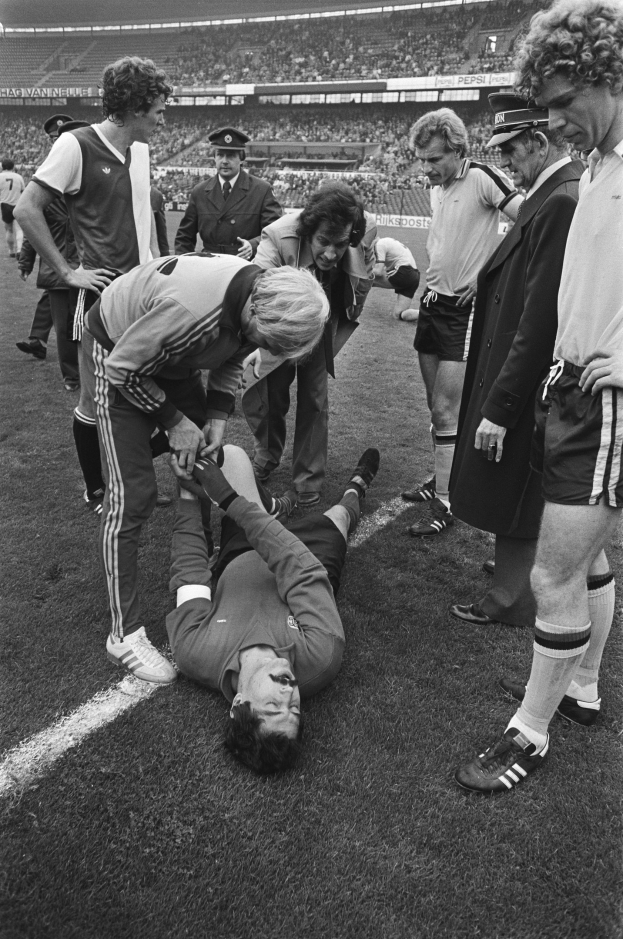 Schwarzes und weißes Bild eines Fußballspielers, der auf dem Boden liegt und von einer Gruppe von Männern und einer Stadionmenge umgeben ist und verletzt zu sein scheint.