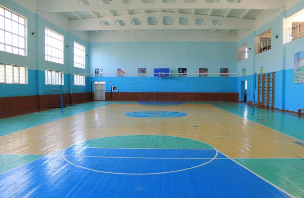 Großer Turnsaal mit einem blauen und weißen Basketballfeld, einem Netz an Stangen, einer Tür, Fenstern, Plakaten an den Wänden, einem Geländer und einem Dach.