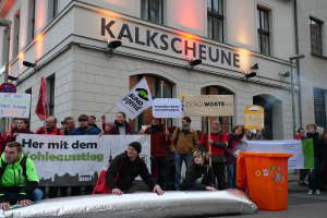 Eine Gruppe von Menschen steht vor einem Gebäude mit Schildern und Plakaten, zwei Personen sitzen im Vordergrund und ein Müllcontainer auf der rechten Seite, während einer Demonstration in Deutschland, mit Gebäuden, Fenstern, Lichtern und Schildern im Hintergrund.