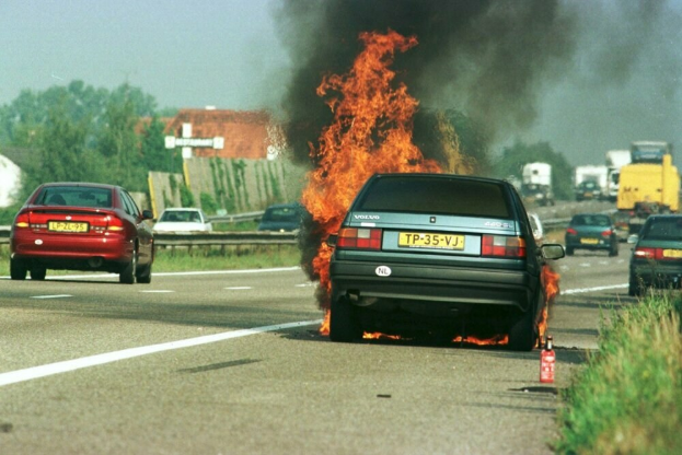 Ein Auto in Flammen auf dem Seitenstreifen, umgeben von anderen Fahrzeugen, mit Bäumen, Gebäuden und einem klaren blauen Himmel im Hintergrund und Gras und einem Feuerlöscher auf der rechten Seite.