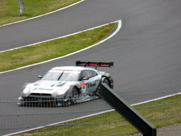 Ein Audi R8 LMS GT3-Rennwagen ist auf der Nürburgring 24-Stunden-Rennstrecke positioniert, umgeben von einem Metallzaun und grünem Gras, mit der Strecke, die sich vor ihm erstreckt.