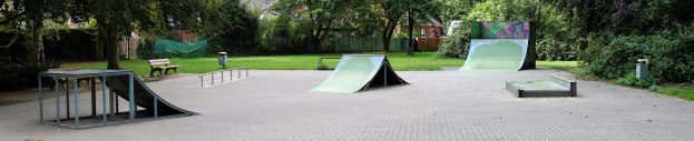 Ein Park mit einer zentralen Skater-Rampe, umgeben von Bänken, Geländern, Grünflächen und Gebäuden im Hintergrund.