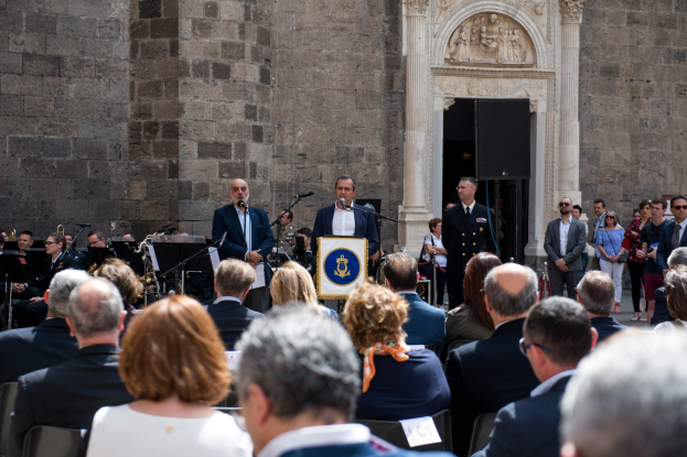 Eine Gruppe von Menschen vor einem Militärkapellenpublikum mit Mikrofonen, einem Podium und einer verzierten Wand im Hintergrund.