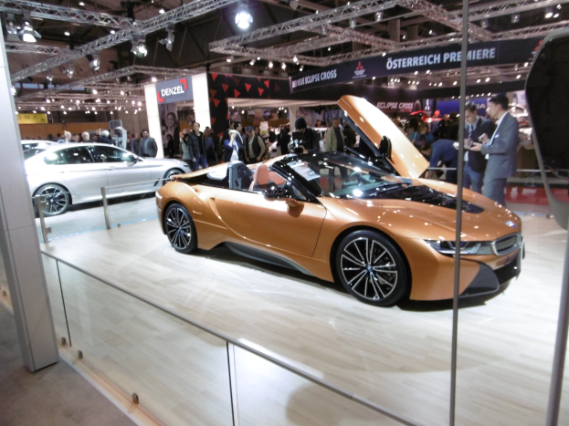 Ein stylischer BMW i8 Roadster mit glänzender schwarzer Lackierung und Chromakzenten, der auf der IAA in Frankfurt präsentiert wird und von formell gekleideten Besuchern umgeben ist.
