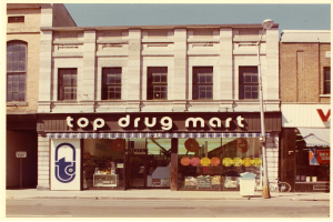 Außenansicht eines Top-Drug-Mart-Geschäfts an einer Straßenecke, das ein Gebäude mit Fenstern, ein Schild, einen Straßenpfahl, ein geparktes Fahrrad, einen Müllcontainer und einen bewölkten Himmel zeigt.
