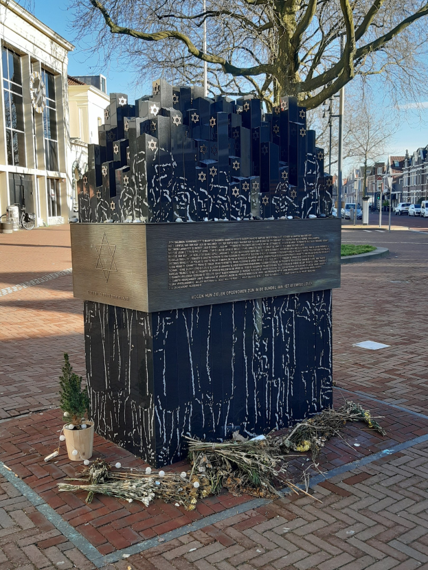 Denkmal in einem Stadtplatz für Holocaust-Opfer, mit einem Baum davor, einer Topfpflanze auf dem Boden, verstreuten trockenen Blättern und im Hintergrund Gebäude, Fahrzeuge, Mäste und Bäume.