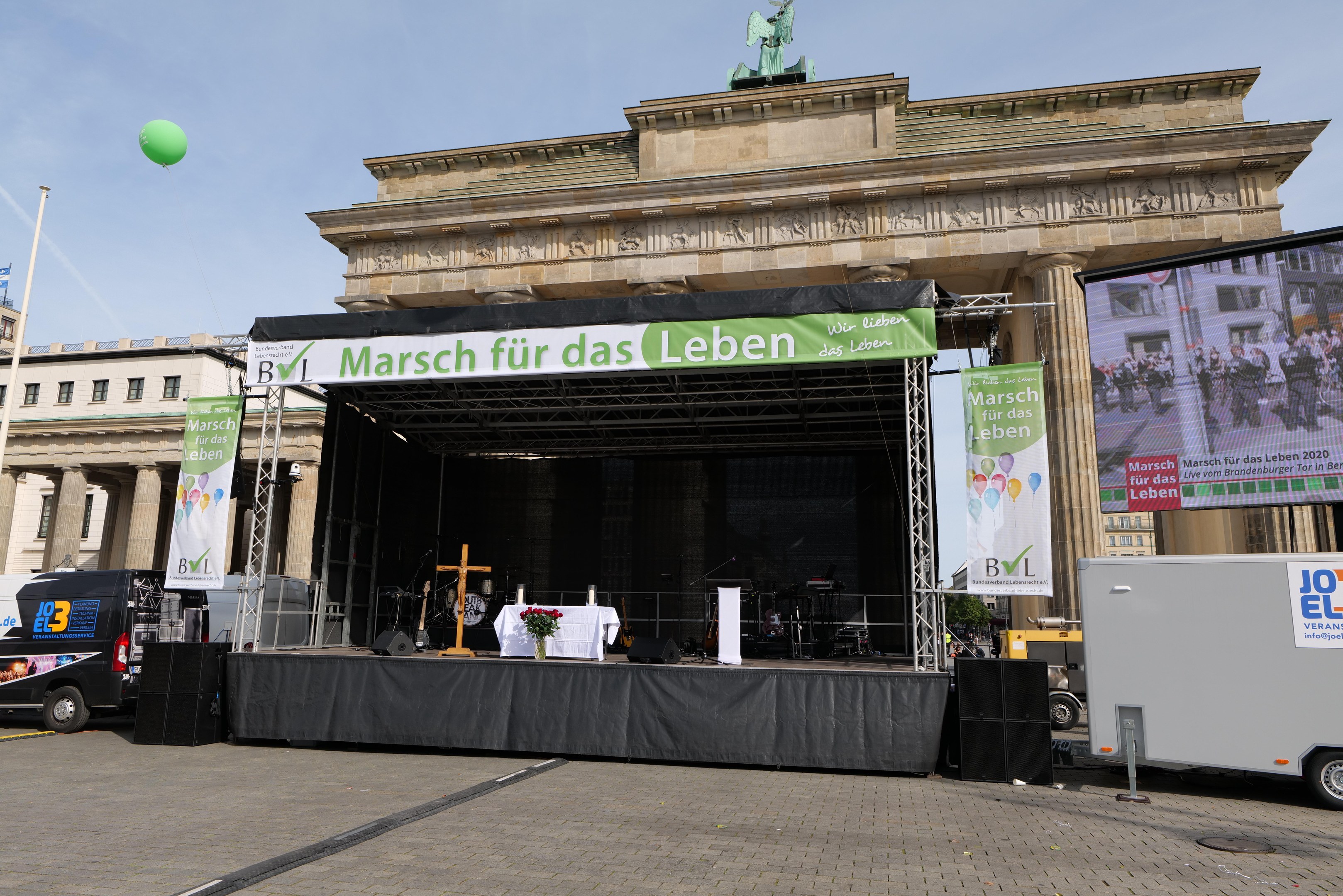 Bühne vor dem Brandenburger Tor mit einem Tisch, der eine Blumenvase, ein Kreuzsymbol und Banner enthält, begleitet von Rednern und Fahrzeugen, mit Gebäuden, einer Statue, Fahnenmast und bewölktem Himmel im Hintergrund.