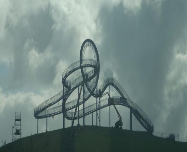 Eine Achterbahn auf dem Höhepunkt eines Hügels unter einem bewölkten Himmel, mit tragenden Pfählen und Strukturen im Hintergrund.