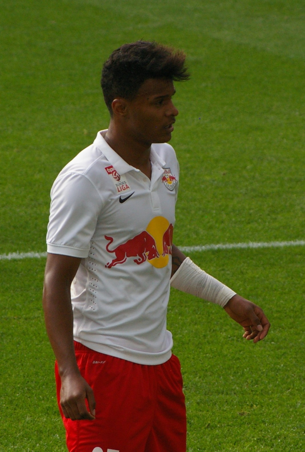 Ein Mann in einem weißen Hemd und roten Shorts steht auf einem Fußballfeld und schaut rechts mit entschlossener Miene, wobei er wie ein Flügelspieler aussieht.