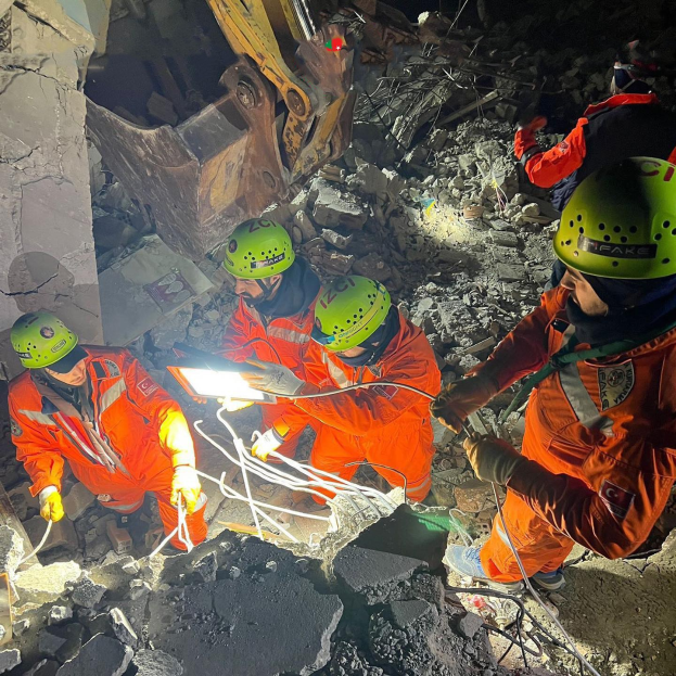 Eine Gruppe von Menschen in orangen Uniformen und grünen Helmen bei der Arbeit an Schutt mit einem Kran im Hintergrund, hölden Werkzeuge und Materialien während einer Rettungsaktion.