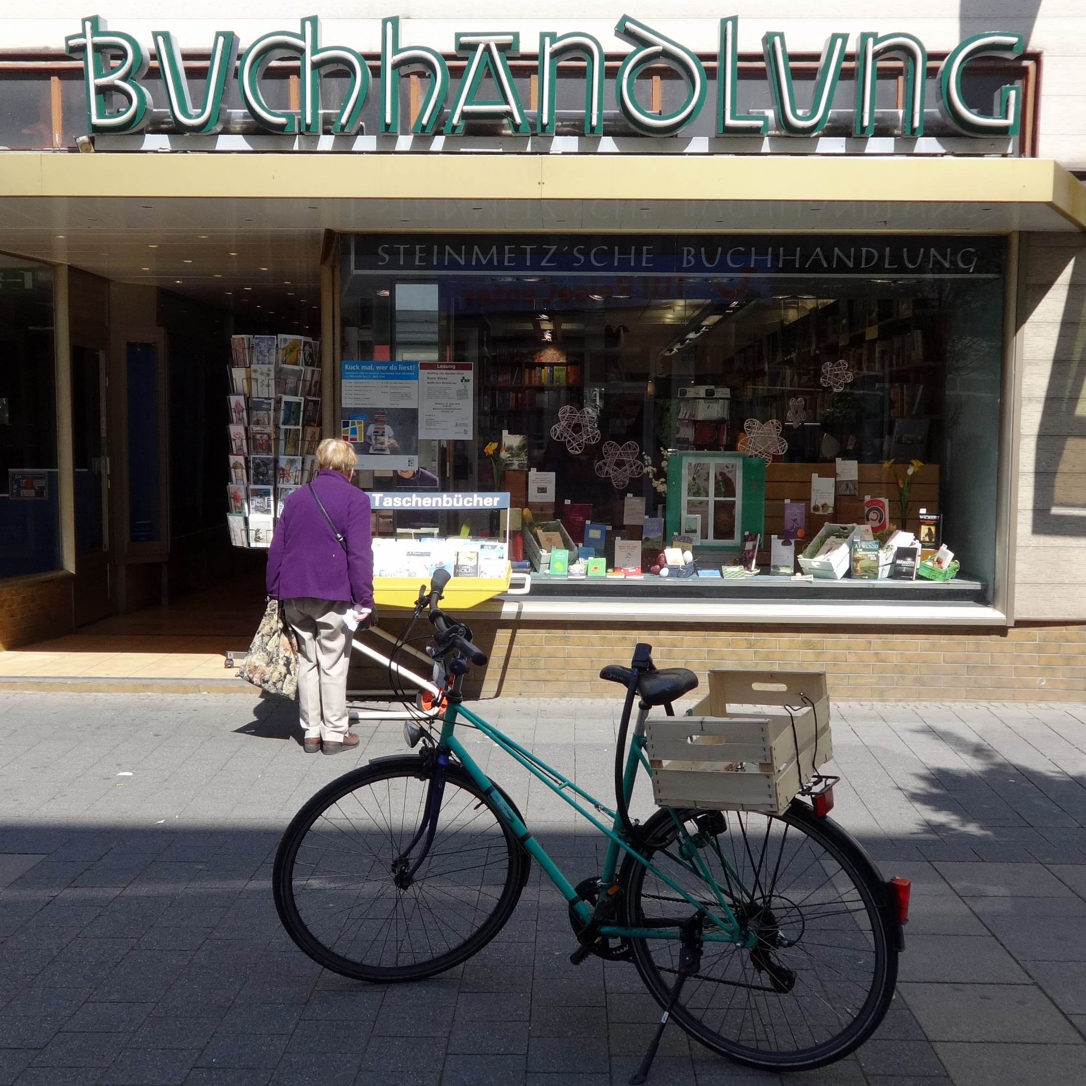 Eine Frau steht neben einem Fahrrad, das vor einem Geschäft geparkt ist, mit einem Fahrradrad in der Vorderansicht und Büchern, die durch die Glasscheibe des Geschäfts sichtbar sind.