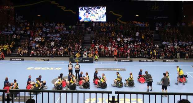 Eine Gruppe von Rollstuhlfahrern bei den Invictus Games Düsseldorf 2020 auf einem Court, umgeben von einem Geländer, mit einer großen Menge im Hintergrund und Stufen, die zu einem Bildschirm führen.