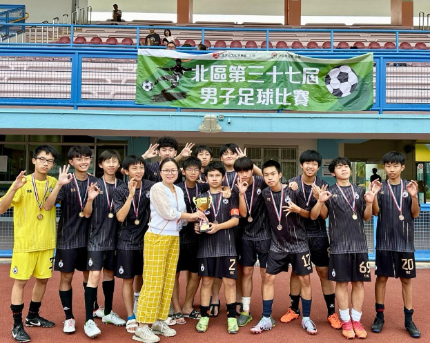 Gruppe junger Männer in Fußballtrikots auf einem Feld stehend, mit Medaillen, einen Pokal haltend, mit einem Banner im Hintergrund beschriftet mit "Yokohama U-16 Jungenfußballmannschaft" und einigen sitzenden Zuschauern.