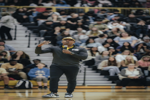 Ein Mann in einer Mütze hält ein Mikrofon auf einem Basketballfeld, hinter ihm sitzen Menschen auf einer Treppe und Taschen liegen auf dem Boden.