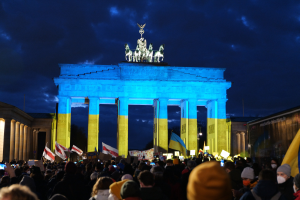 Eine Menschenmenge mit Fahnen und Plakaten steht vor dem Brandenburger Tor in Berlin, Deutschland, mit einer Fahne auf der rechten Seite und bewölktem Himmel.