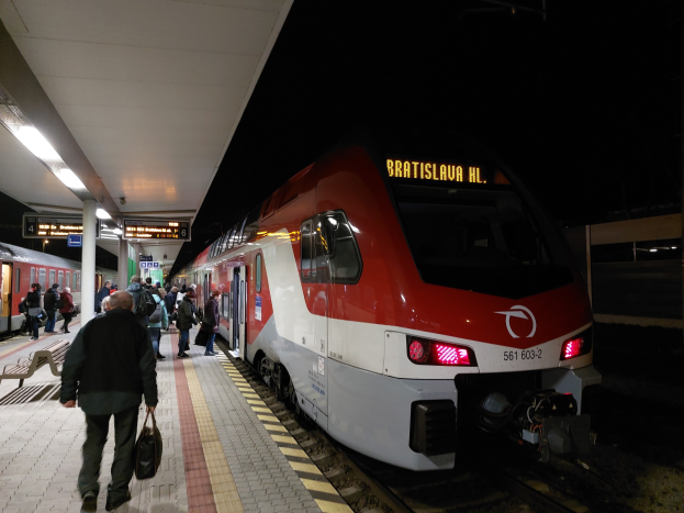 Roter und weißer Zug hält nachts auf einem Bahnhof mit Wartenden auf dem Bahnsteig und beleuchteten Bänken links.