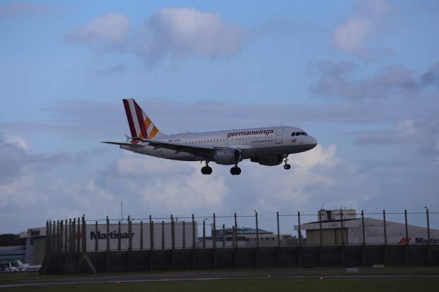 Germanwings Airbus A320-200 beim Start am Frankfurter Flughafen, mit Gras darunter, einem Zaun im Vordergrund, Gebäuden und Bäumen im Hintergrund und einer bewölkten Himmel