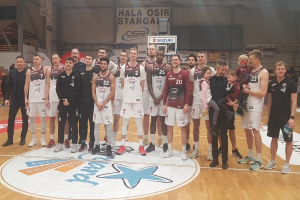 Männer- und Kinder-Gruppe auf einem Basketballfeld mit Schildern "Hala Osir Starga", sitzende Zuschauer, ein Basketballkorb, Metallstangen, Deckenleuchten und eine Wand im Hintergrund.