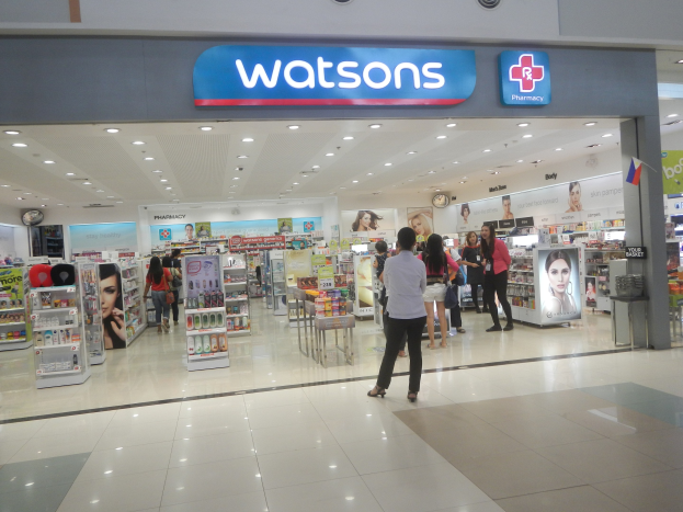 Außenansicht einer Watsons-Apotheke in einem Einkaufszentrum mit Menschen, Produkten, Regalen, Beschriftung, einer Wanduhr und Deckenbeleuchtung.