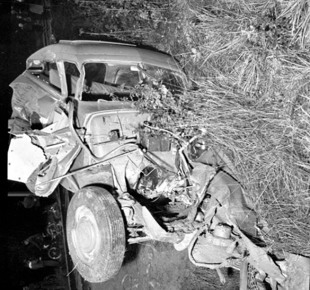Ein Schwarz-Weiß-Foto eines schwer beschädigten Autos am Straßenrand mit einer eingedrückten Front, zersplitterten Fenstern, einem Baum im Hintergrund und verstreuten Gegenständen um die Unfallstelle.