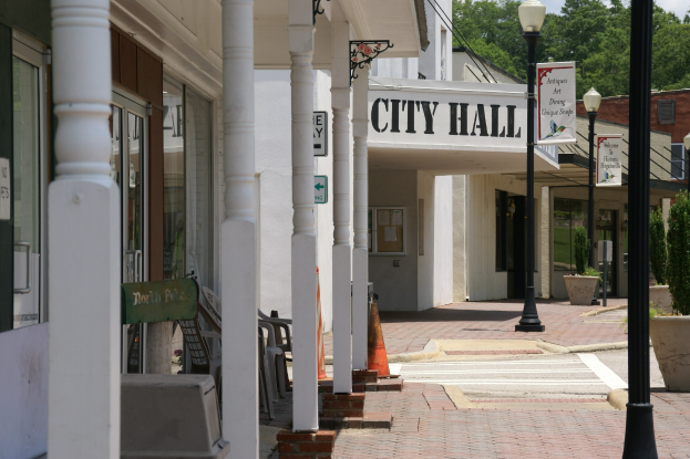 Ein belebter Stadtplatz mit Menschen auf Stühlen sitzend und stehend, umgeben von Topfpflanzen, Metallabsperrungen, Schildern, Straßenlaternen mit Flaggen, Gebäuden und einem bewölkten Himmel.