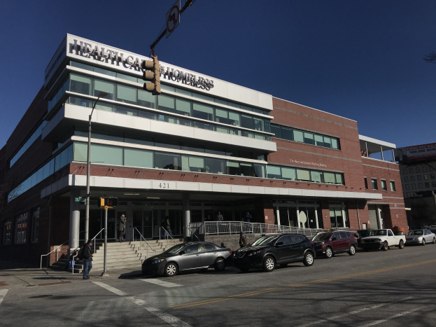 Großes Gebäude mit der Aufschrift "New York City Health Care Homeless Center" mit geparkten Autos, Passanten auf einem Gehweg, Straßenmöbel und Bäumen unter einem klaren blauen Himmel.