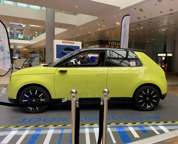 Gelbes wasserstoffbetriebenes Elektroauto vor einem Gebäude mit Absperrpollern, Bannern, Menschen, Schildern, einer Topfpflanze, Deckenleuchten und Lüftern.