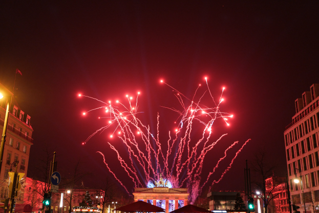 Eine belebte Stadtstraße an einem Silvesterabend in Berlin, mit Gebäuden, Bäumen, Laternenmasten, Verkehrszeichen, Zeltplanen, Menschen und einem prächtigen Feuerwerk am Himmel.