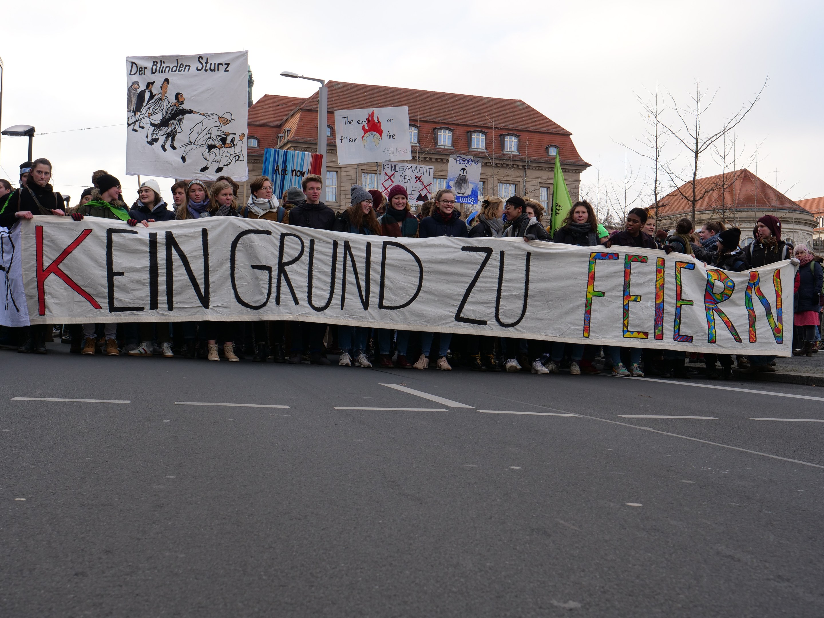 Protestierende mit einem Banner mit der Aufschrift 'Kein Grund zu Feiern' gegen deutsche Sparmaßnahmen, mit Straßeninfrastruktur und Gebäuden im Hintergrund bei klarem Himmel.