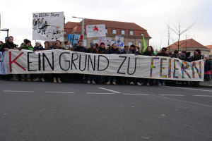 Protestierende mit einem Banner mit der Aufschrift 'Kein Grund zu Feiern' gegen deutsche Sparmaßnahmen, mit Straßeninfrastruktur und Gebäuden im Hintergrund bei klarem Himmel.
