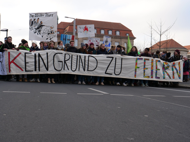 Protestierende mit einem Banner mit der Aufschrift 'Kein Grund zu Feiern' gegen deutsche Sparmaßnahmen, mit Straßeninfrastruktur und Gebäuden im Hintergrund bei klarem Himmel.