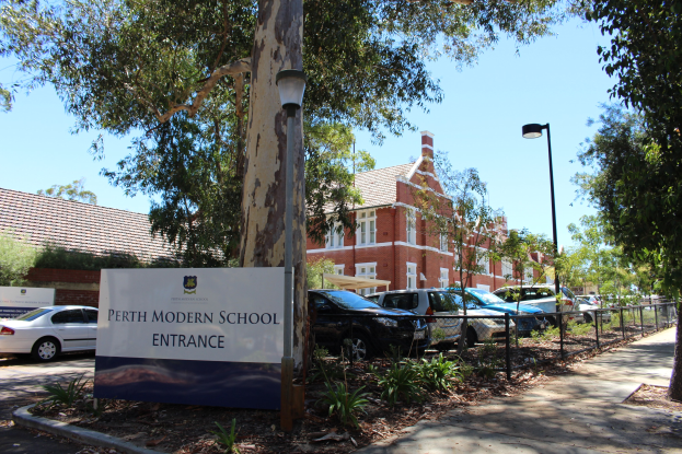 Einfahrt zur Perth Modern School mit einem Schild, Bäumen, Pflanzen, einem Zaun, einem Straßenpfosten, parkenden Autos, Gebäuden mit Fenstern und einem bewölkten Himmel.