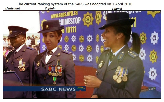 Gruppe von Menschen in Uniformen und Hüten vor einem Banner mit Text oben und unten, der "Das aktuelle Rangsystem der SAP wurde am 1. April 2010 übernommen" lautet.