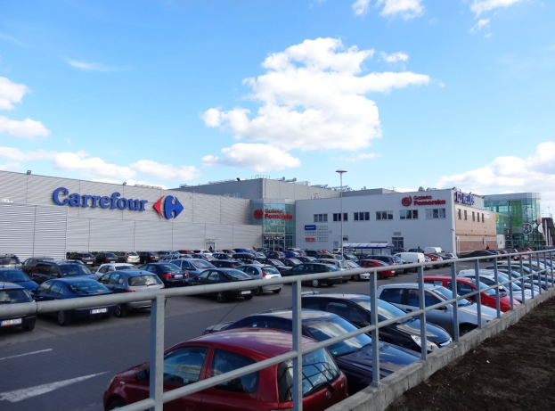 Großes Autohaus mit mehreren geparkten Fahrzeugen, umgeben von städtischer Infrastruktur einschließlich Gebäuden, Straßenlaternen und Fahrzeugen auf der Straße, unter einem bewölkten Himmel.