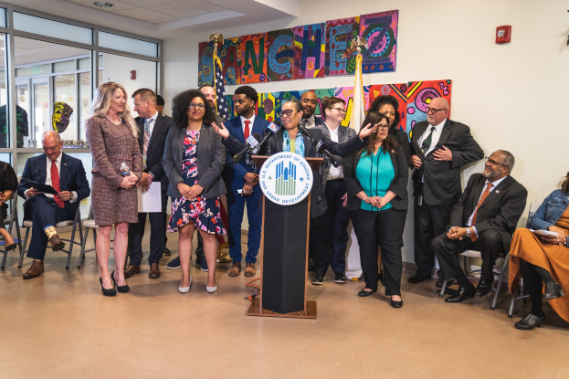 Gruppe von Menschen vor einem Rednerpult mit Mikrofon und einer Tafel mit der Aufschrift "Sancho-Stadtrat tritt dem Stadtrat bei", mit sitzenden und stehenden Zuhörern, Fahnen, einem Feuerlöscher und Deckenleuchten im Hintergrund.