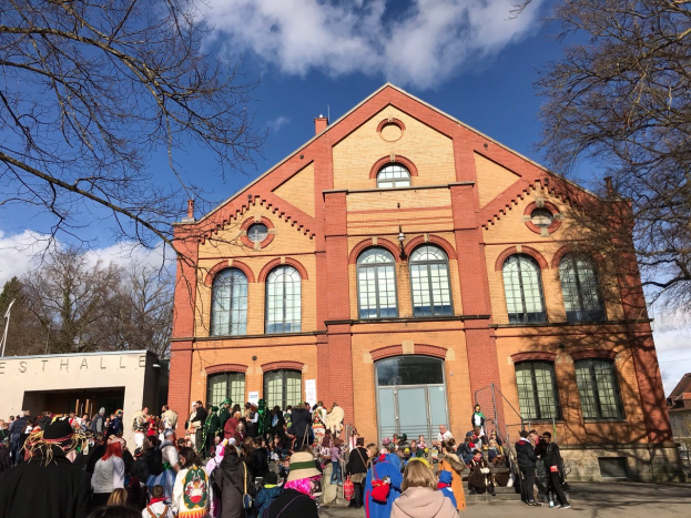 Eine große Gruppe von Menschen steht vor einem mehrstöckigen Gebäude mit Fenstern und Türen, umgeben von Bäumen unter einem bewölkten Himmel, einige tragen Mützen und tragen Taschen, was auf ein Stadtfest hinweist.
