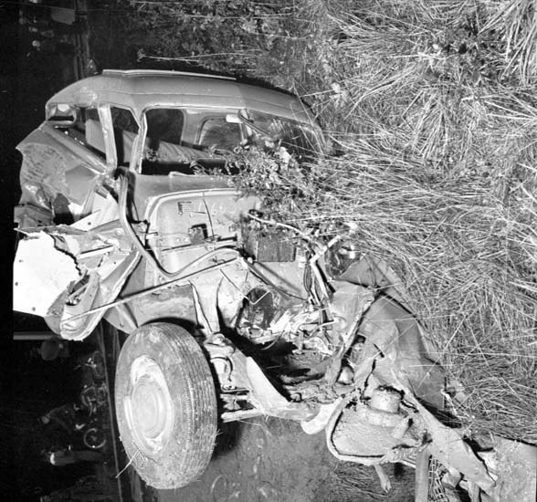 Ein Schwarz-Weiß-Foto eines schwer beschädigten Autos am Straßenrand, mit eingedellter Front, zersplitterten Fenstern, einem Baum im Hintergrund und verstreuten Gegenständen an der Unfallstelle.