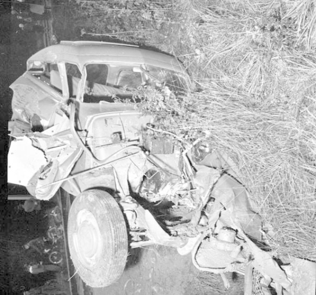 Ein Schwarz-Weiß-Foto eines schwer beschädigten Autos am Straßenrand, mit eingedellter Front, zersplitterten Fenstern, einem Baum im Hintergrund und verstreuten Gegenständen an der Unfallstelle.