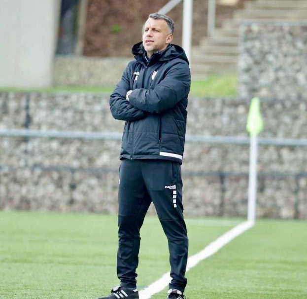 Ein Mann in Schwarzjacke und -hose steht auf einem Fuballfeld mit überkreuzten Armen, der als Trainer während einer Trainingseinheit erscheint.