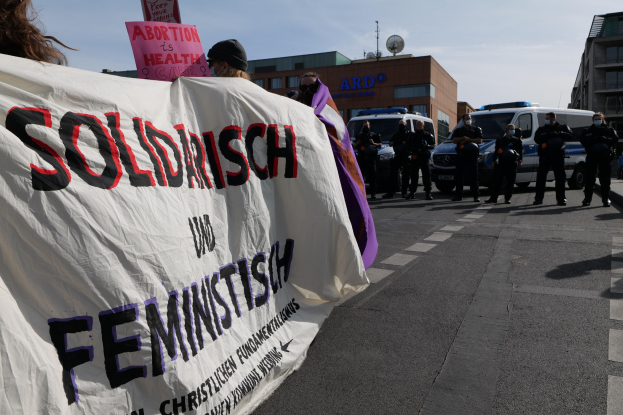 Eine Gruppe von Menschen geht eine Straße entlang und hält eine 'Solidarität und Feminismus'-Schleife in der Hand, mit parkenden Fahrzeugen, Gebäuden, einer Schüsselantenne und einem klaren blauen Himmel im Hintergrund.