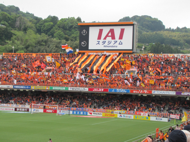 Fußballspiel in einem Stadion mit einer großen Zuschauermenge, grünem Rasen, Torstangen, Fahnen, Großbildschirm, Bäumen und einem klaren blauen Himmel.
