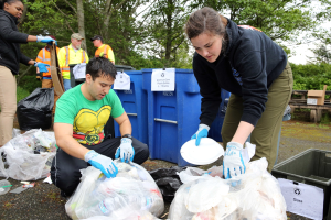 Eine Gruppe von Menschen, die Müll in einem Park sammeln, mit zwei Personen in der Mitte, die Handschuhe tragen und Teller halten, umgeben von weggeworfenen Gegenständen, einem Mülleimer, einer hölzernen Bank, Bäumen und einem klaren blauen Himmel.