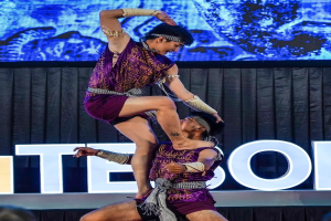 Ein Mann und eine Frau in lila Kleidern führen Akrobatik auf der Bühne durch, wobei der Mann die Frau in seinen Armen hält, während das Publikum unten zusieht und ein schwarzer Vorhang und ein Bildschirm im Hintergrund zu sehen sind.