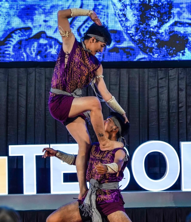 Ein Mann und eine Frau in lila Kleidern führen Akrobatik auf der Bühne durch, wobei der Mann die Frau in seinen Armen hält, während das Publikum unten zusieht und ein schwarzer Vorhang und ein Bildschirm im Hintergrund zu sehen sind.