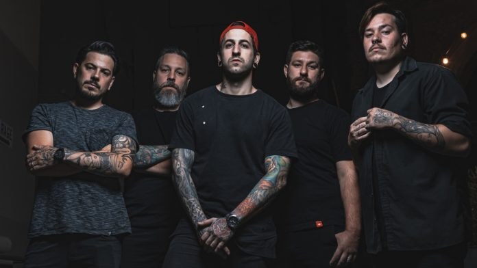 Eine Gruppe von Männern, die in einem schwach beleuchteten Raum stehen, von rechts beleuchtet, während sie offenbar ein Interview für Avenged Sevenfold geben.